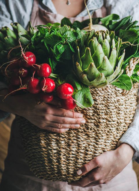 Female farmer in linen apron holding basket of fresh garden vegetables and greens in her hands, rustic wooden barn wall at background, horizontal composition. Local market or organic produce concept